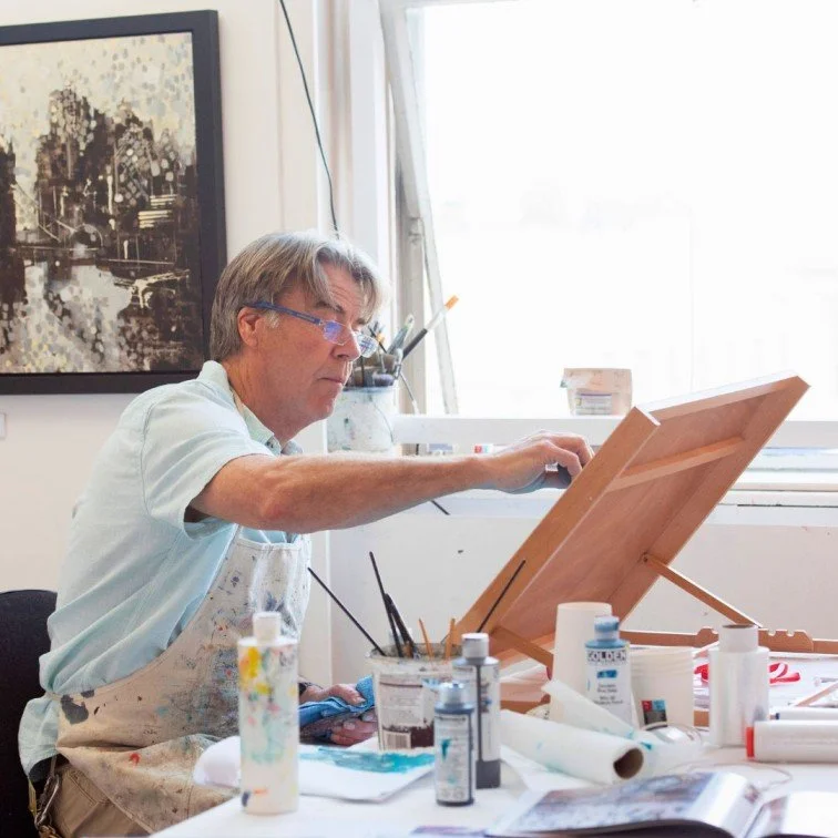 One landscape painter is inside his art studio painting an artwork from his easel, while surrounded by paints and brushes. There is one original framed painting hanging on the wall behind him.