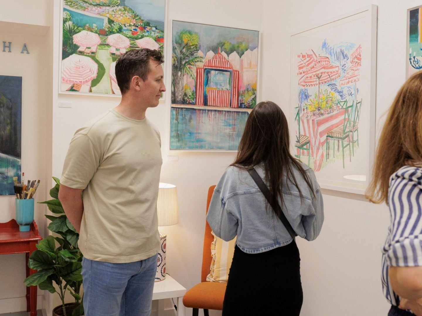 People viewing colorful landscape and abstract paintings in an art gallery.