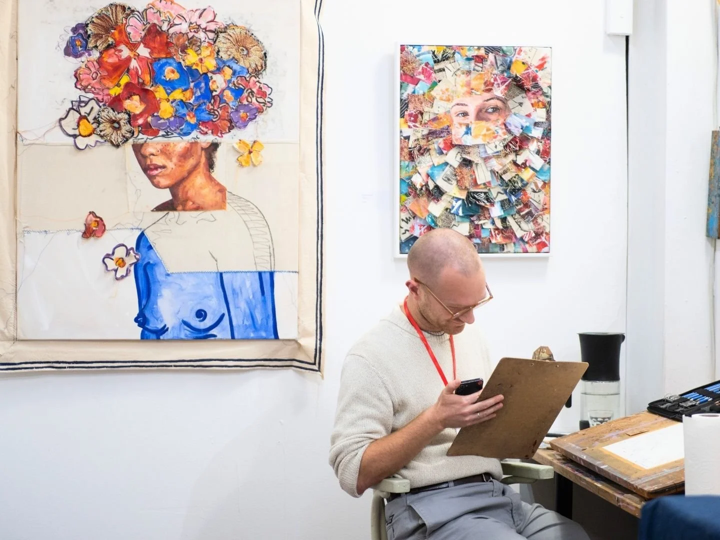 An artist sitting in a studio with two colourful mixed media portraits hung on a white wall behind him. He is looking at a painting paper while seated at a sketching table with art supplies and a water bottle.