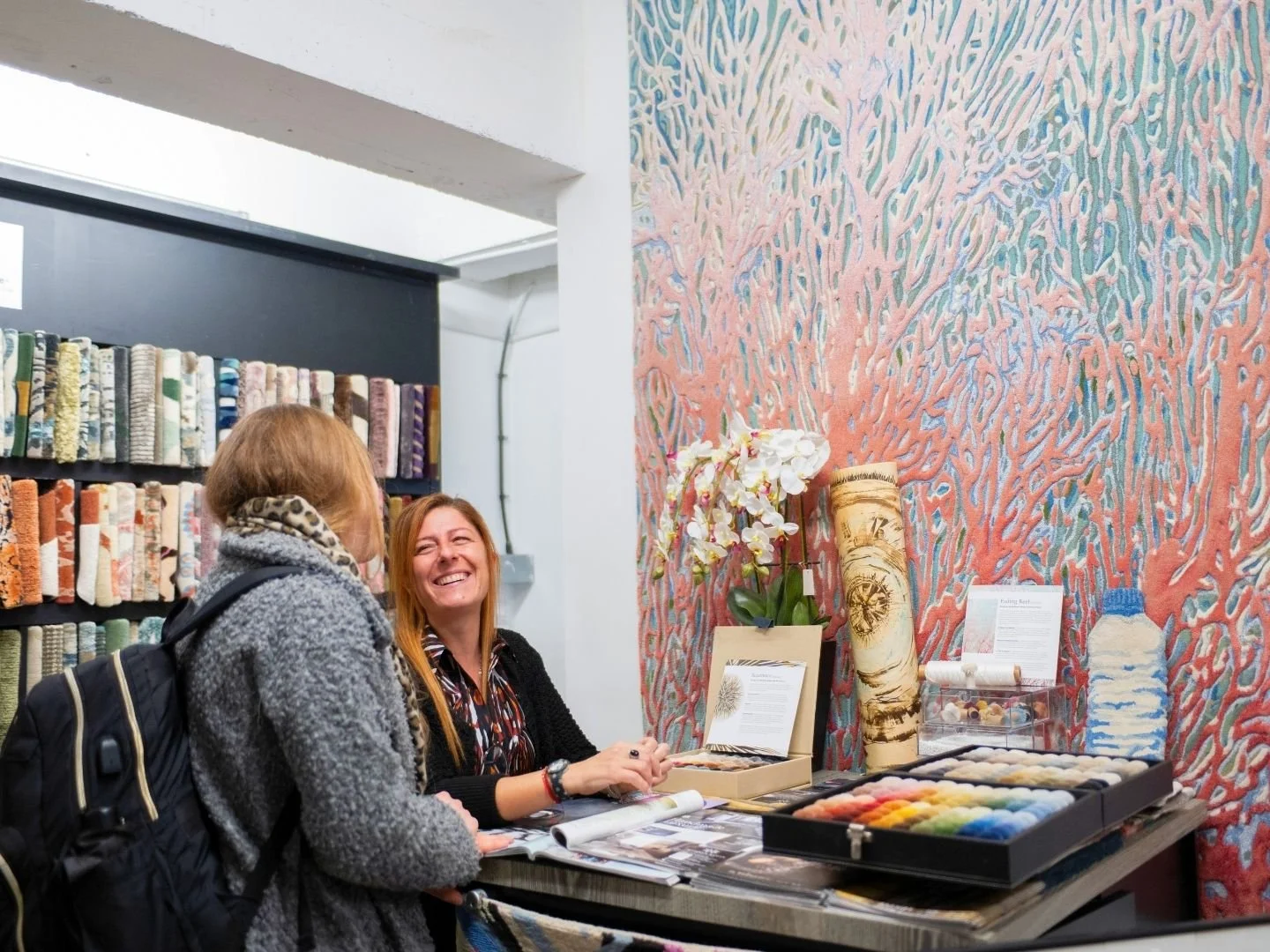 Two women at a fabric or craft store, one with red hair smiling and the other with brown hair, engaging in conversation at a table with fabric and craft supplies, colorful wall with textured patterns and shelves with fabric rolls in the background.