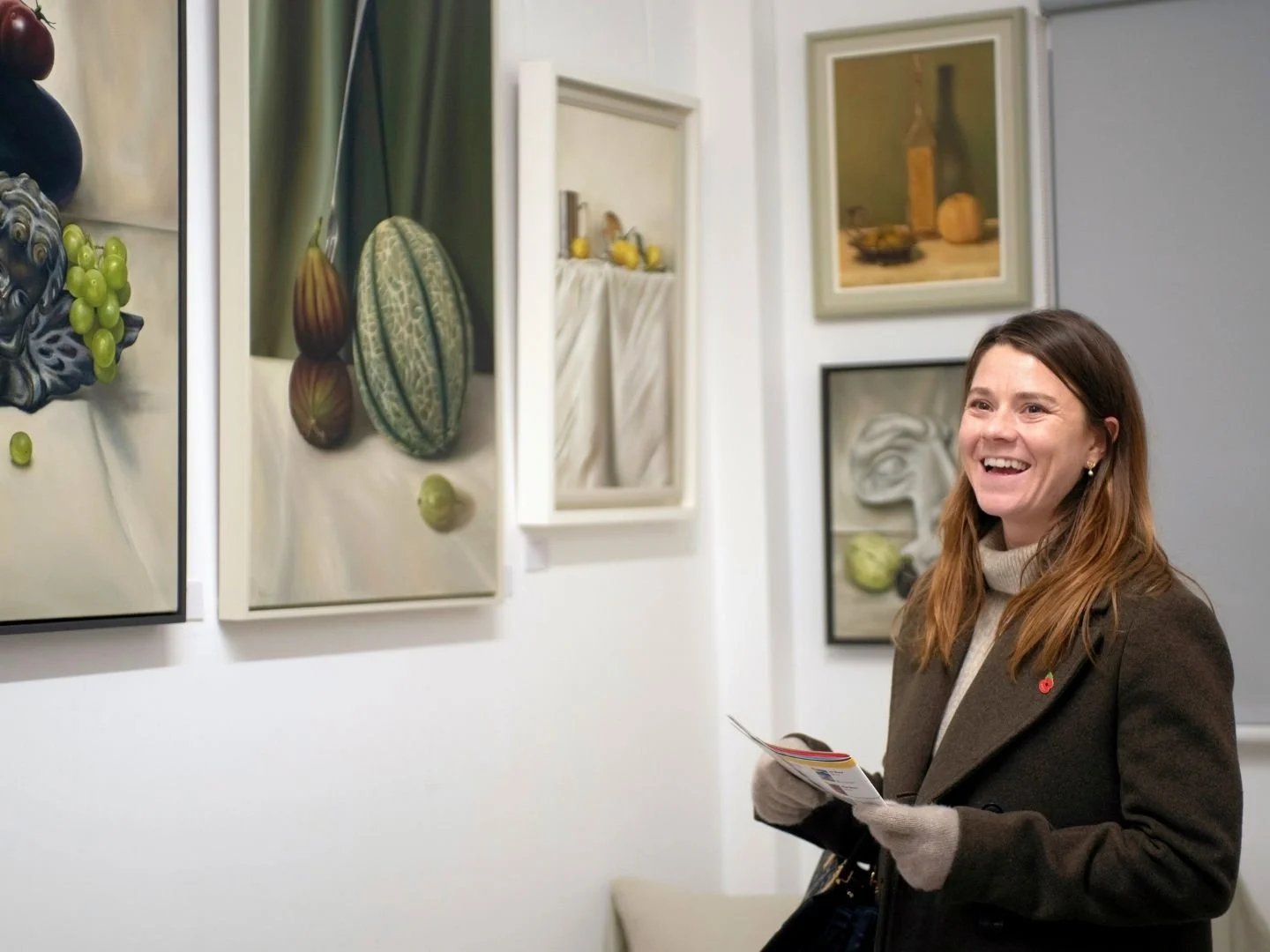 A woman with brown hair, wearing a coat and gloves, is smiling while looking at artwork in an art gallery.