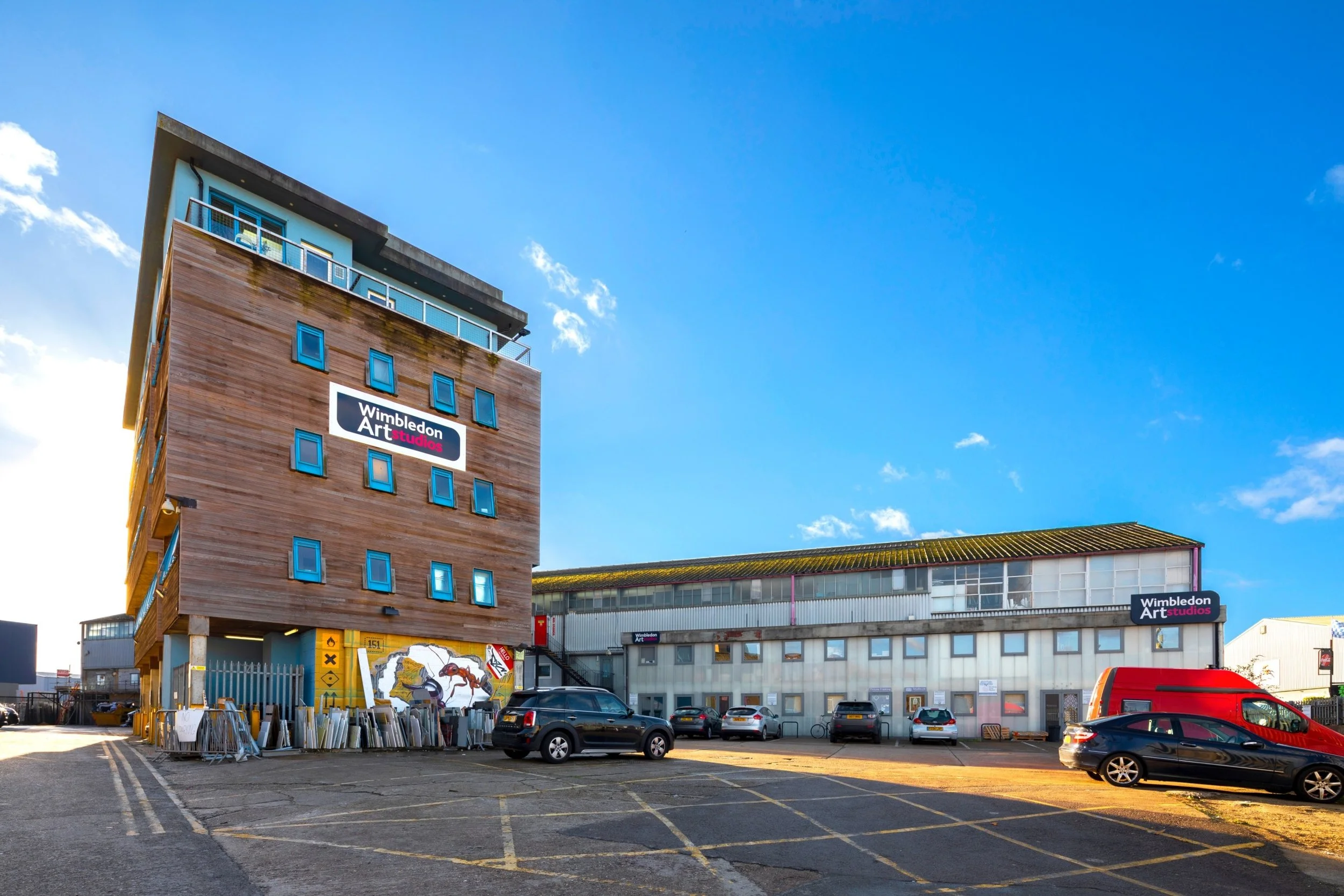 Two buildings from Wimbledon Art Studios with its free car park on a sunny, blue sky day. The Blue Studios wooden building has a graffiti on one of the walls.