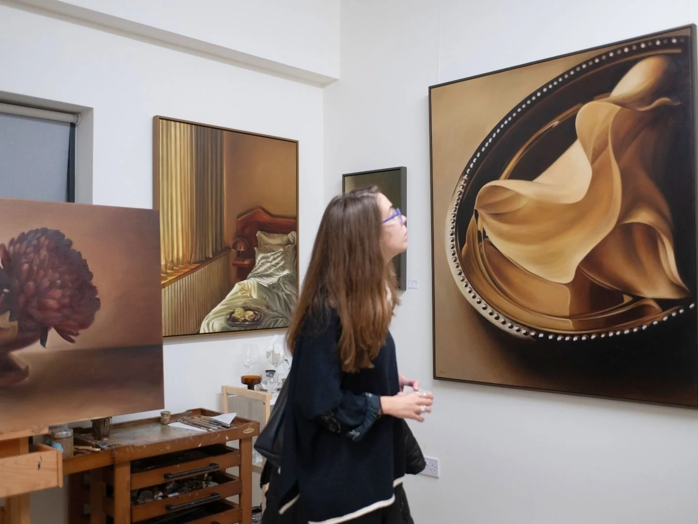 A woman with long hair and glasses looks at a large, abstract painting in an art gallery. The painting features swirling, smooth shapes in shades of brown and gold.