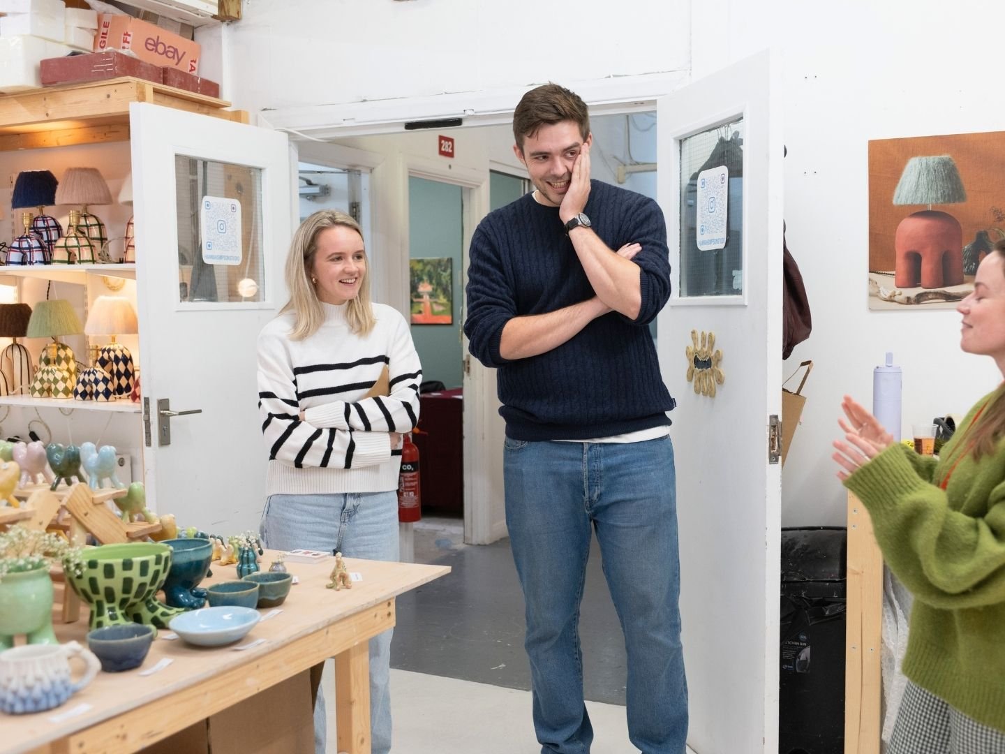 Three people in a pottery shop, two women and a man, smiling and talking. The woman on the left wears a white and black striped sweater, the man is in a navy sweater, and the woman on the right wears a green sweater.