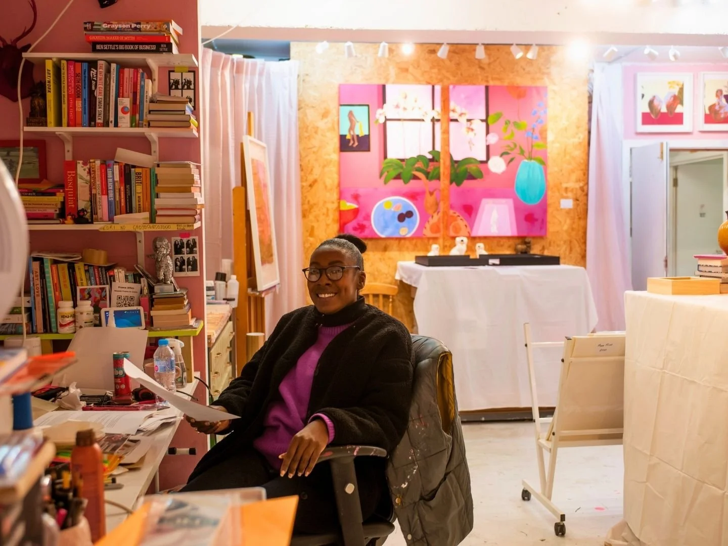 An artist in her art studio with glasses smiling at a desk filled with books, papers, and art materials in her creative space with colourful paintings and artwork on the walls.