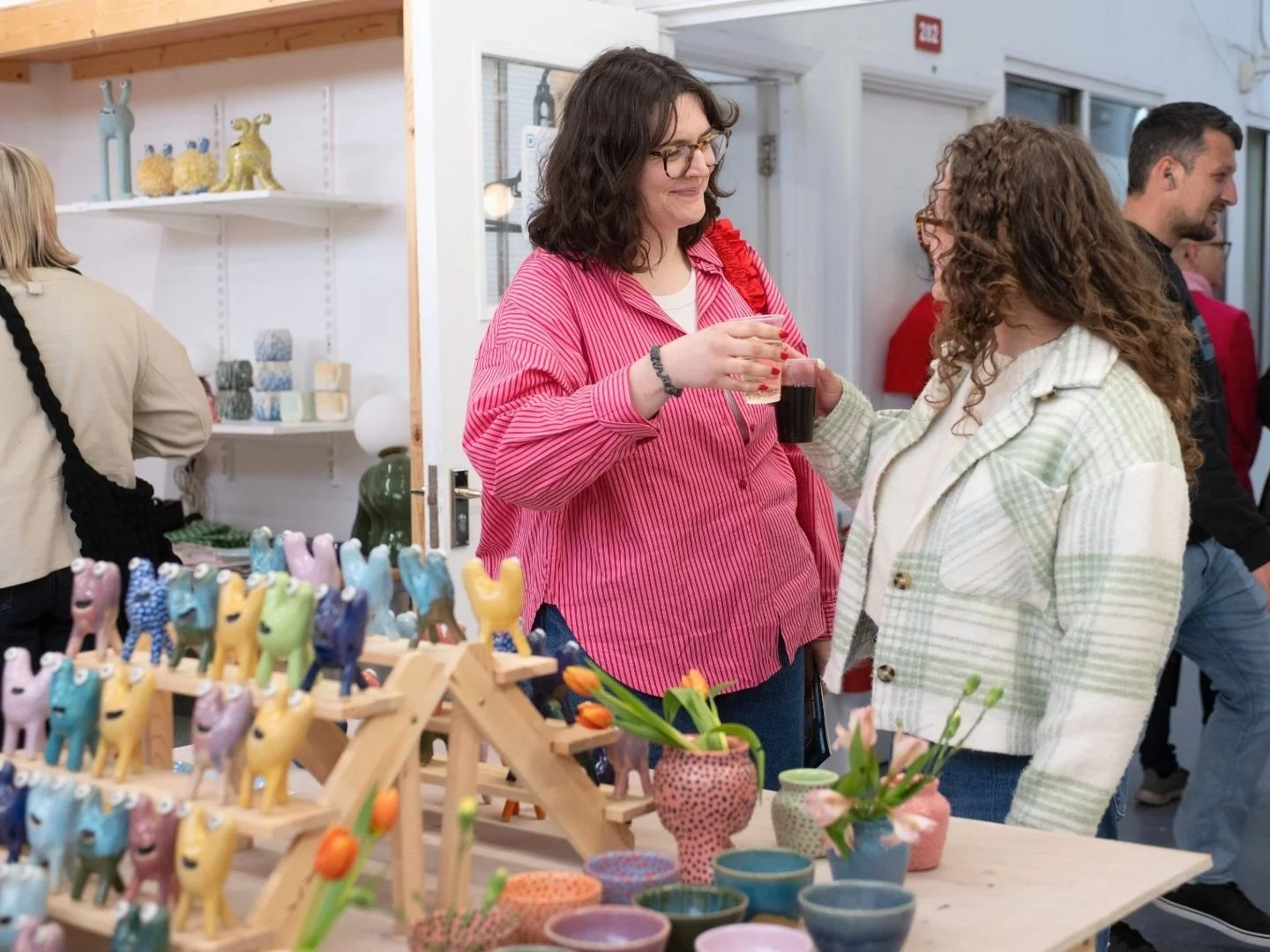 Two women are talking at an art fair, holding drinks, with colourful pottery and figurines on display in front of them.