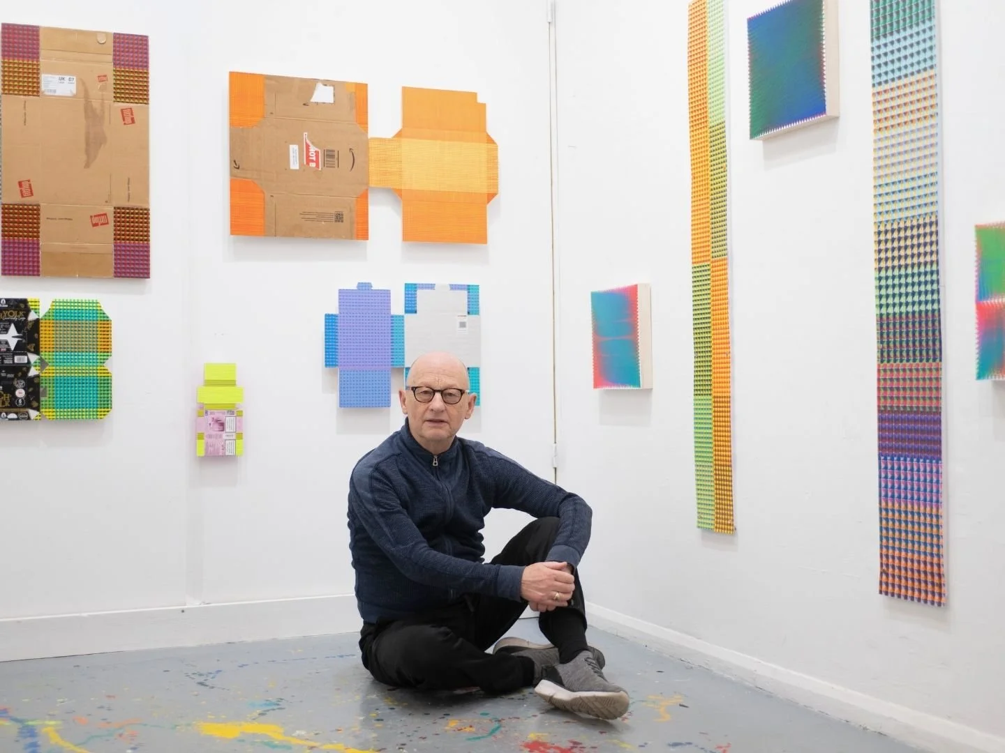 An artist man with glasses sits on the floor in an art studio surrounded by colourful abstract artwork on the white walls.