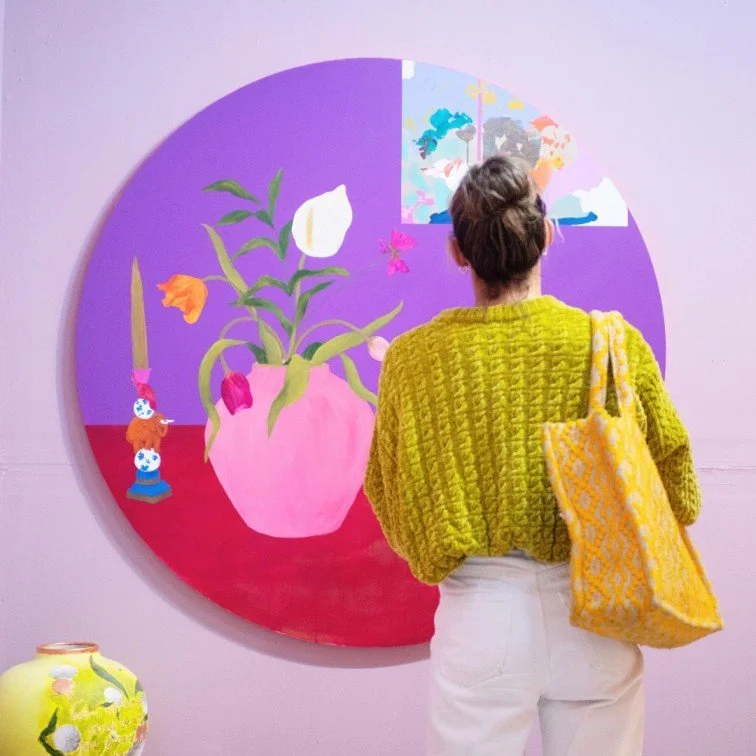 A visitor from Wimbledon Art Fair is standing in front of an oval oil still life painting made by one of the community artists. The walls are painted in purple, and the artwork is also painted in warm colours.