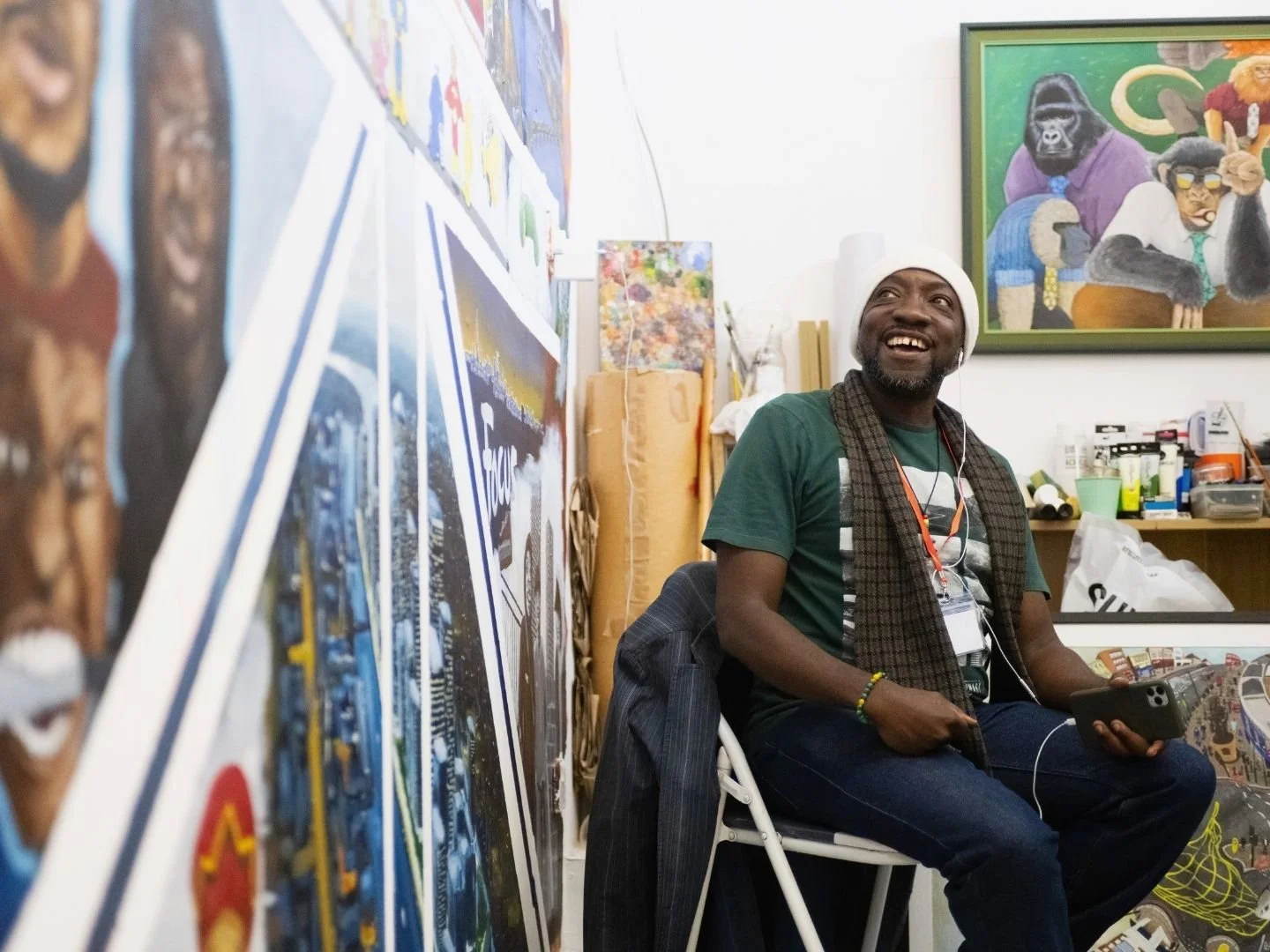 An artist sitting on a white chair in his art studio, smiling and looking upward to a hand painted wall, holding a phone, wearing a white hat, green t-shirt, and a scarf. Behind him are colourful paintings and art supplies.