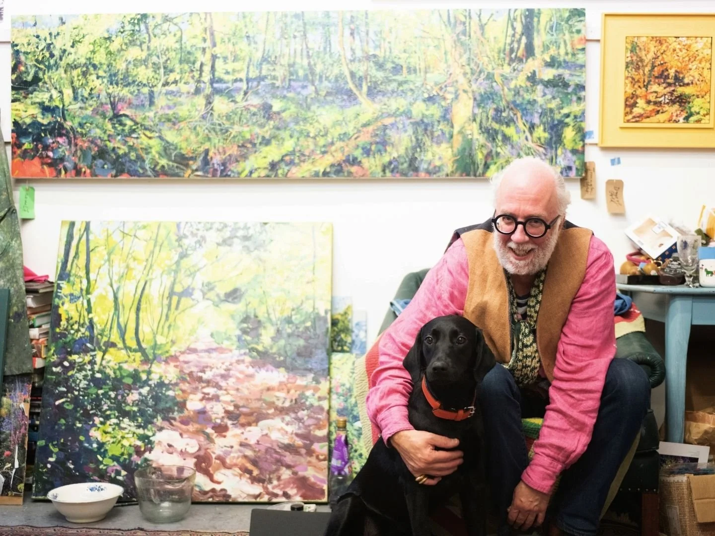 An elderly man with glasses, a beard, and a striped shirt sitting on a chair in an art studio, holding a black dog with an orange collar. Behind them are large landscape paintings of forest scenes.