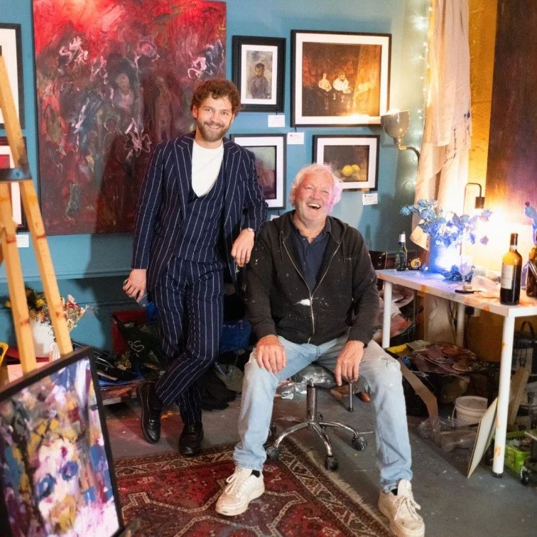 Two community artists are inside of an art studio, surrounded by abstract paintings. Some of the paintings hanging on the wall are framed. There is a table with art materials on top, as well with a bottle of wine for the studio visitors of the show.