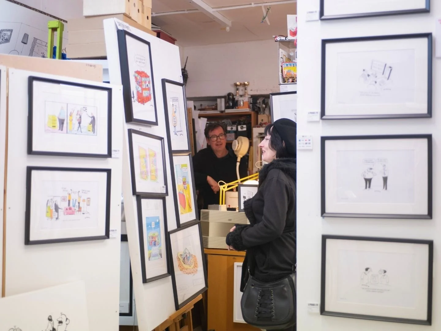 A woman shopping for artwork in an art studio, looking at framed illustrations and cartoons displayed on white partitions, while the cartoonist artist, a man with glasses, discusses with her behind the counter.