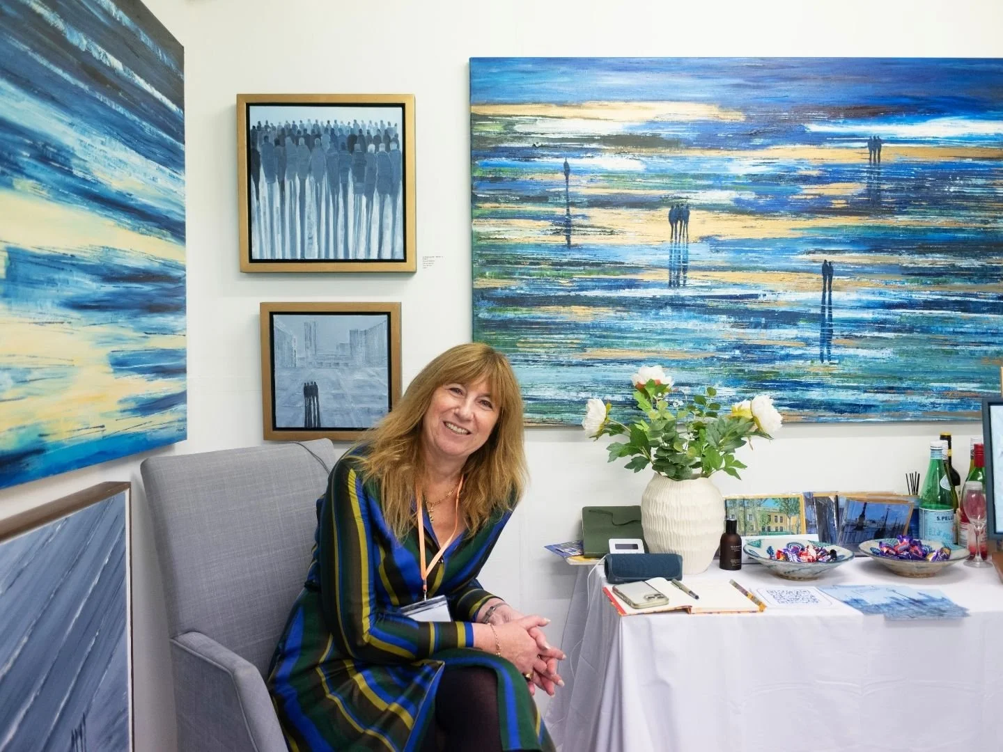 A woman with long blonde hair smiling while sitting on a gray chair in an art studio, surrounded by blue abstract paintings and a table with water bottles, candies, a flower vase, and notebooks.