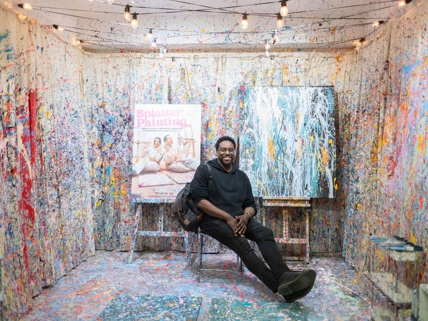 Man smiling and seated in a splatter paint art studio with multicolored paint splashes on the walls, ceiling, and floor, and abstract paintings behind him.