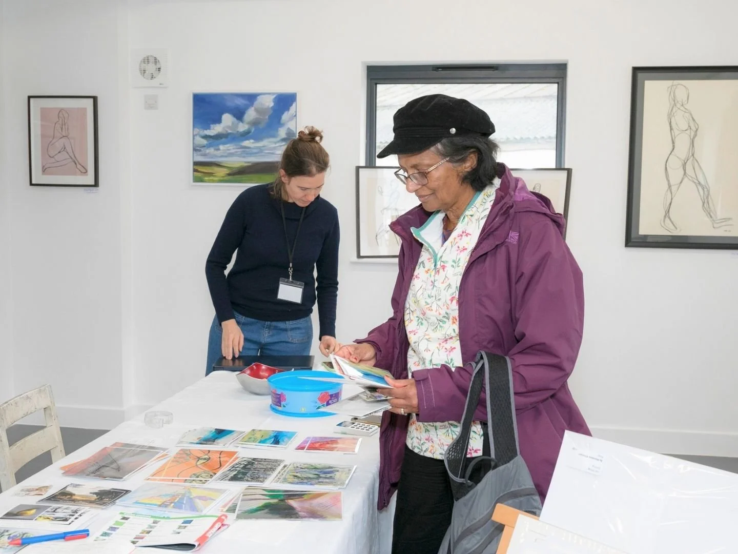 Two women, one elderly and one younger, browsing artwork at an art gallery, with paintings on the wall behind them and framed art on a table.