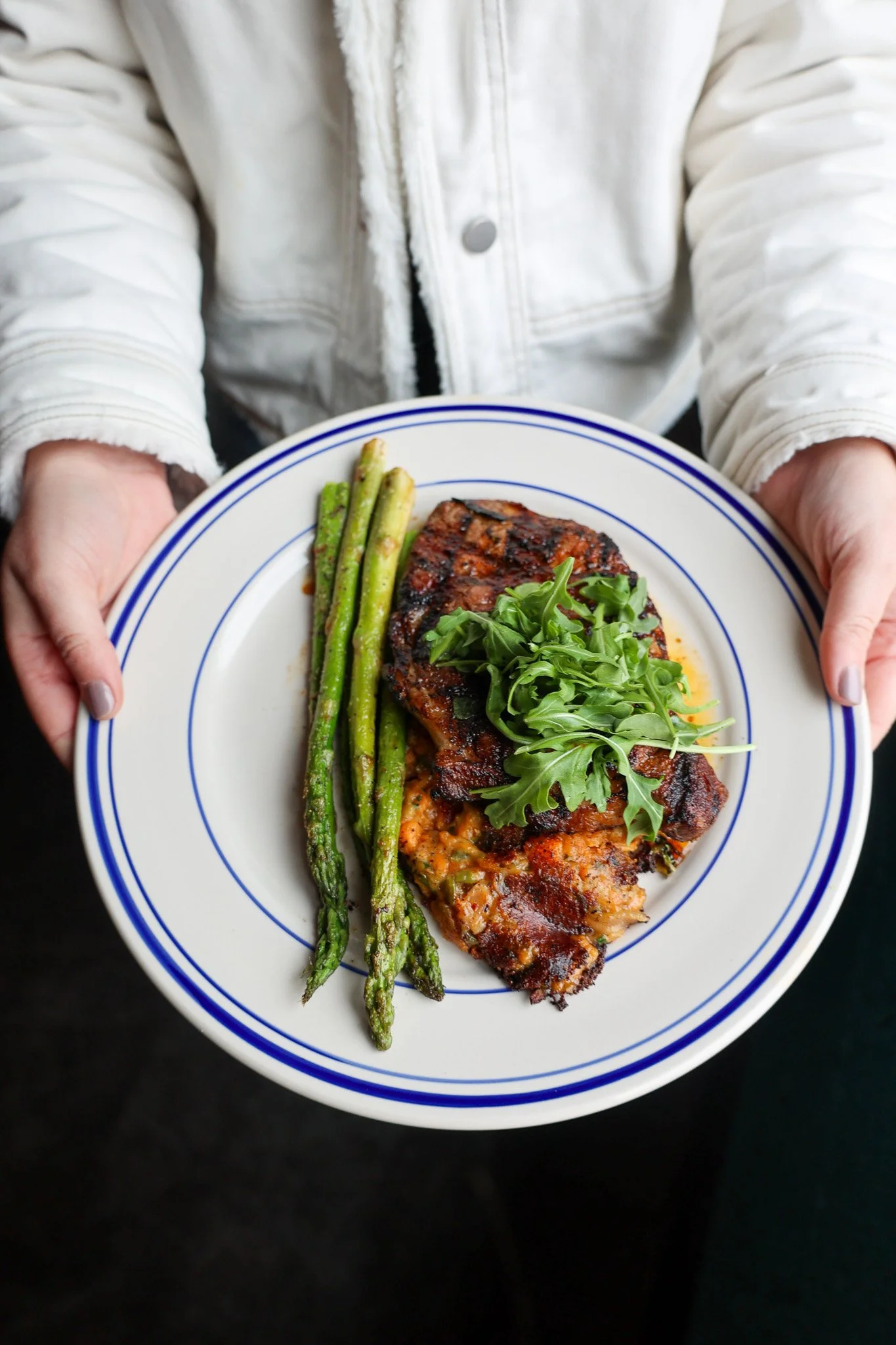 Person holding a white plate with blue rim containing grilled steak, roasted asparagus, and a small pile of arugula.