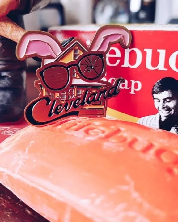 A decorative pin with sunglasses, bunny ears, and the words 'Cleveland' and a house, placed on a surface with pizza in the foreground and a red box in the background.