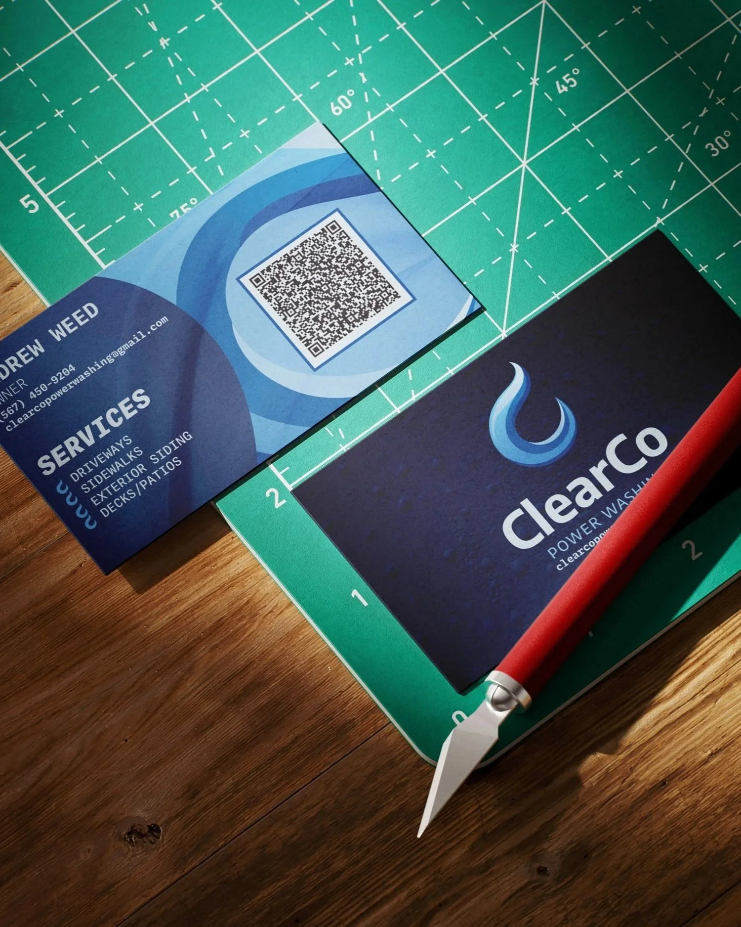 Business card for Andrew Weed with contact information, business services listed, next to a Clearco Power Wash brochure, on a wooden table with a cutting tool.