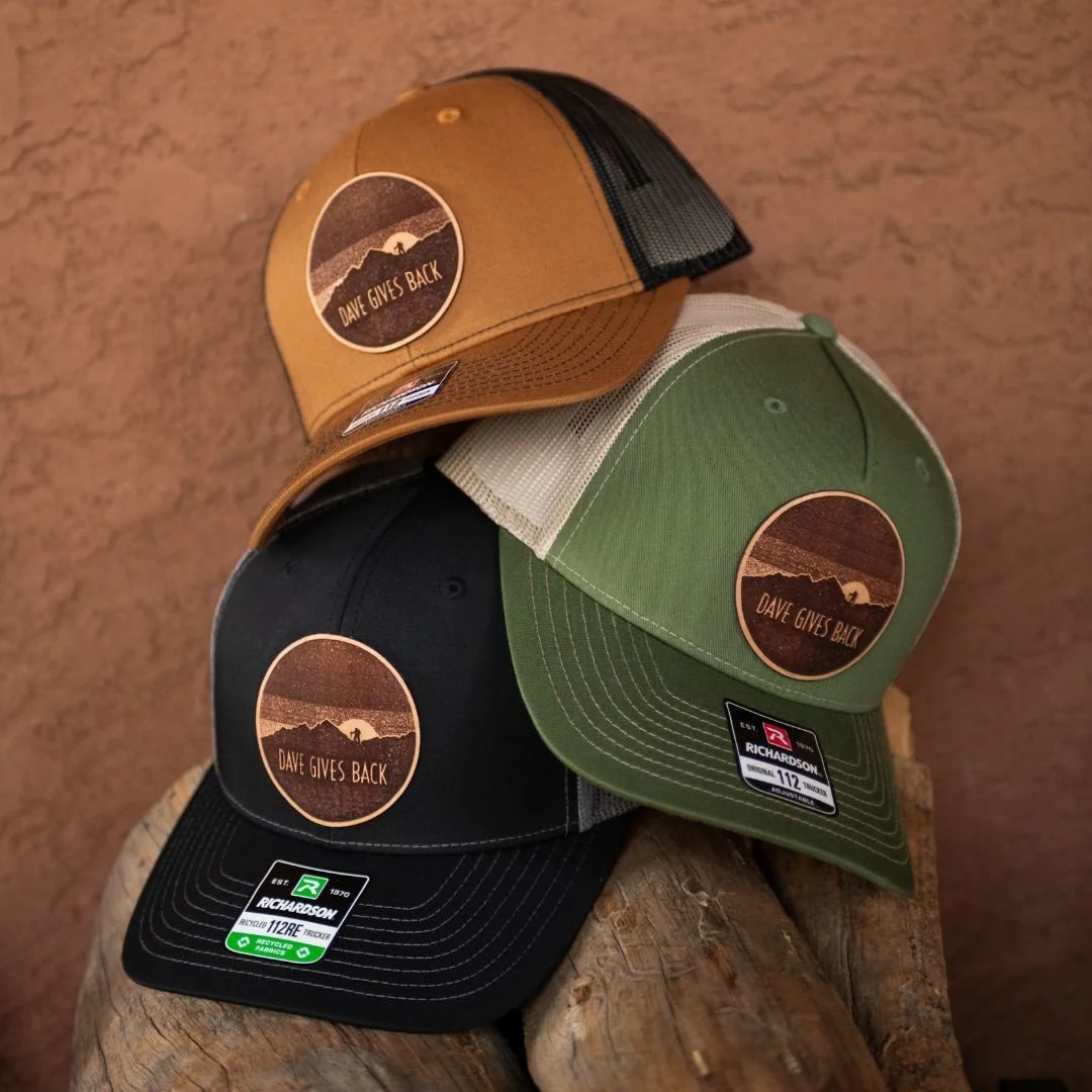 Three baseball caps stacked on a wooden surface, each with a circular patch that says "DAVE GIVES BACK" with a mountain and sunrise graphic, in tan, green, and black colors.
