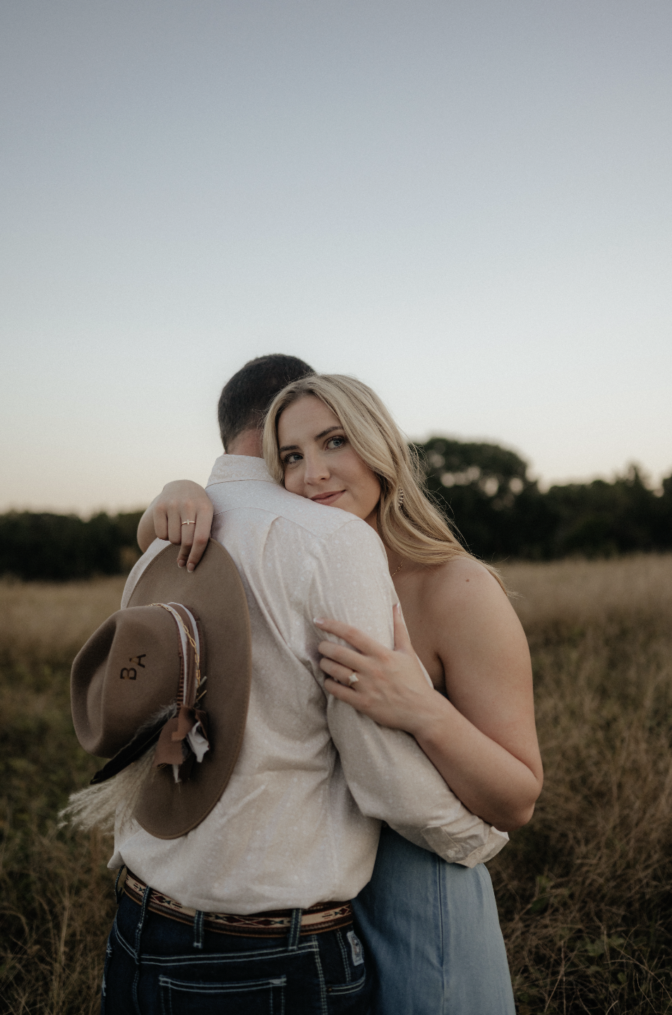 A woman with blonde hair hugging a man in a field during sunset, holding a cowboy hat with the initials 'B A'.