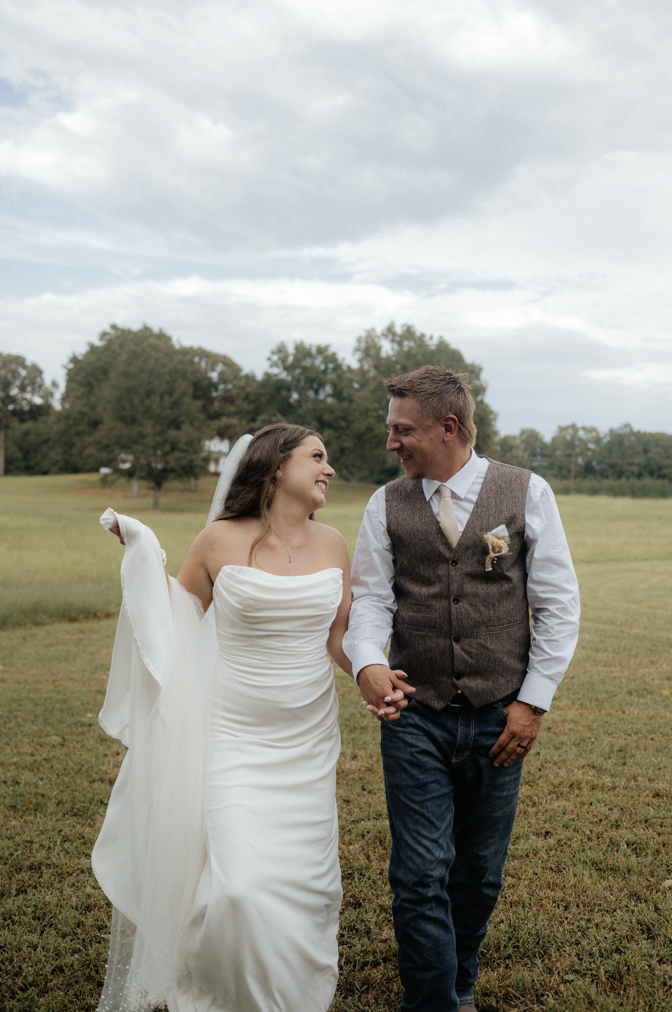 A newlywed couple holding hands and smiling at each other in a grassy field with trees and a cloudy sky in the background.