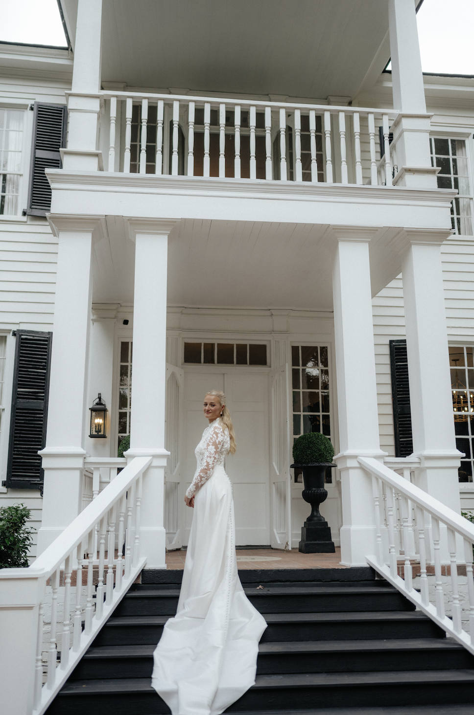 A woman in a white wedding dress standing on the stairs of a white house with a porch, black shutters, and a potted plant.