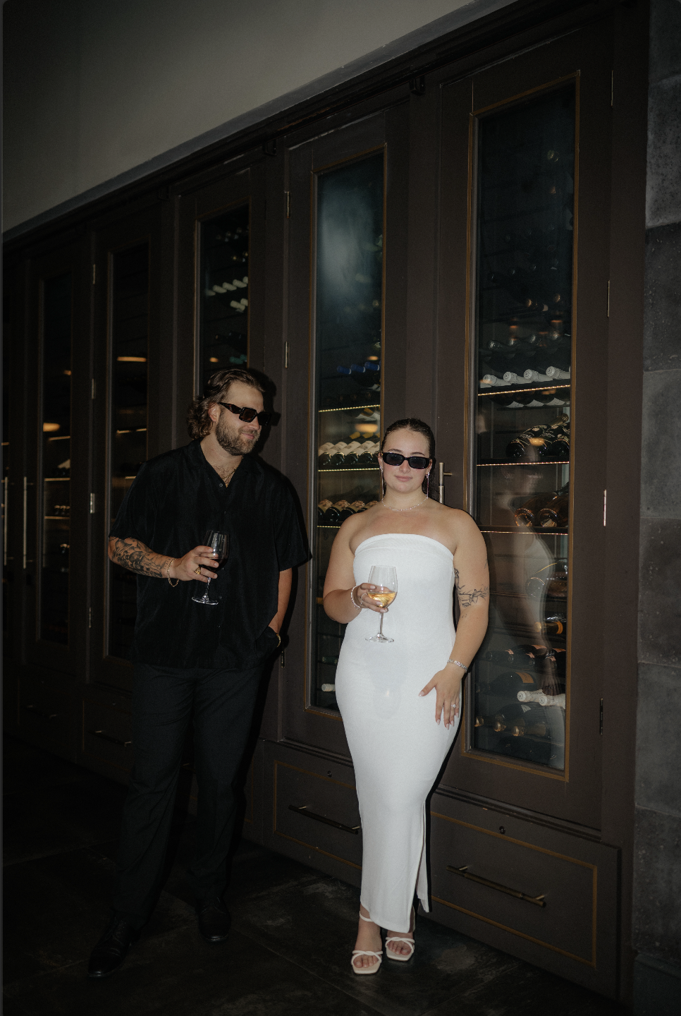 A man and woman wearing sunglasses stand in front of a wine fridge, each holding a glass of wine in a dimly lit setting.