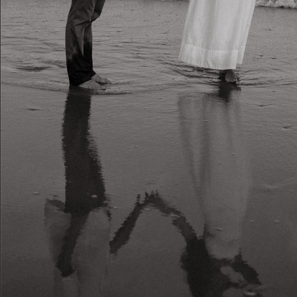 People standing and walking barefoot on wet sand at the beach, with their reflections visible in the water.