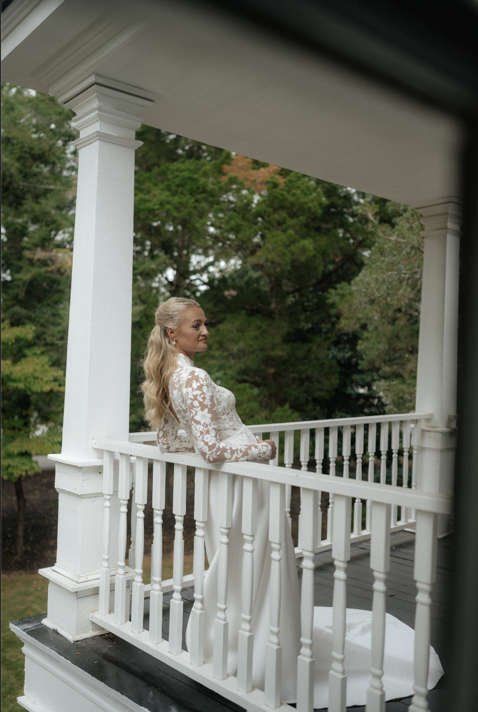 A bride in a white wedding dress with lace top standing on a porch with white railing, looking to the side, surrounded by green trees.