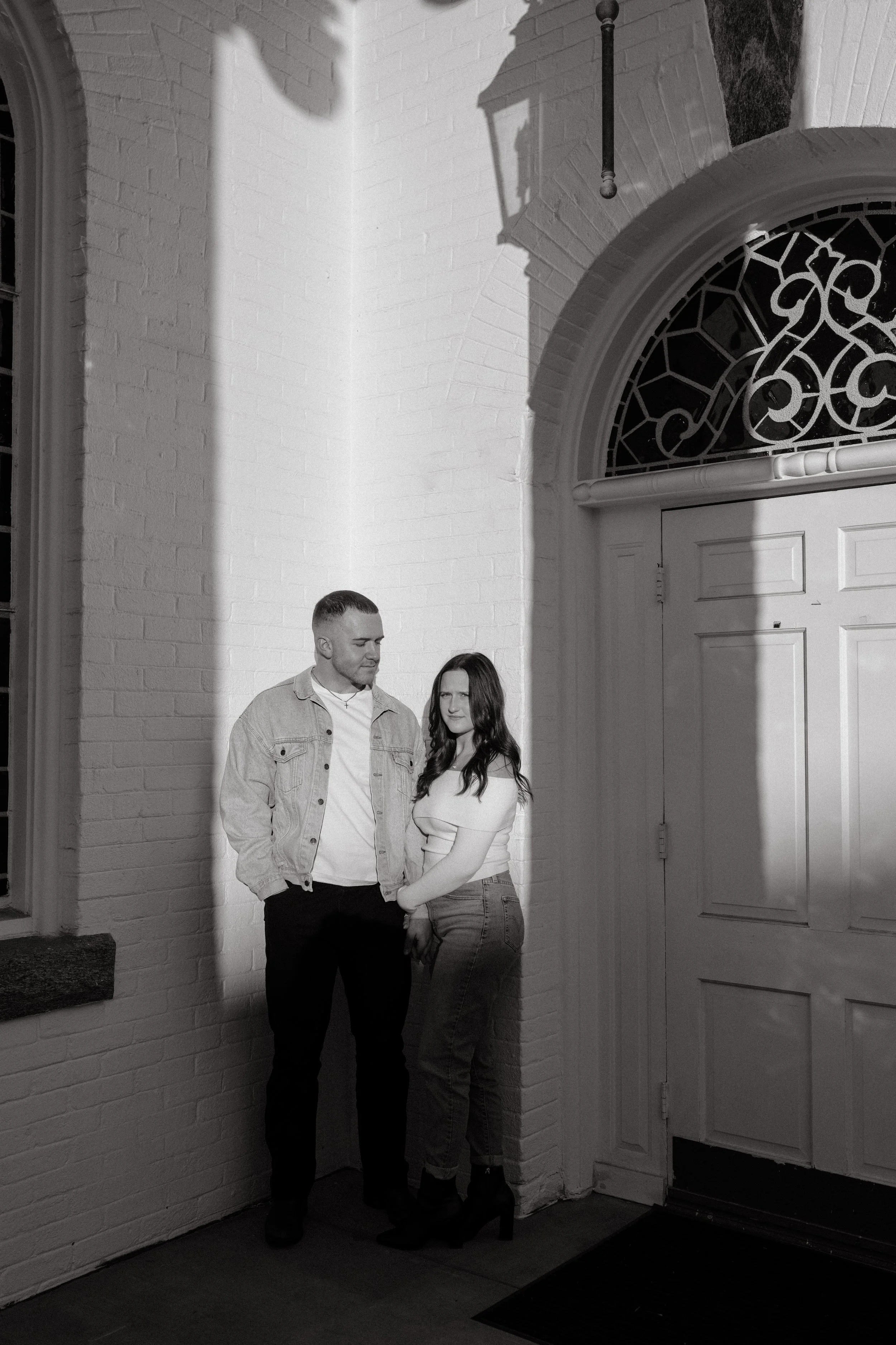 A black and white photo of a man and woman standing close together near a white brick wall and a door with a decorative window, with shadows cast on the wall.
