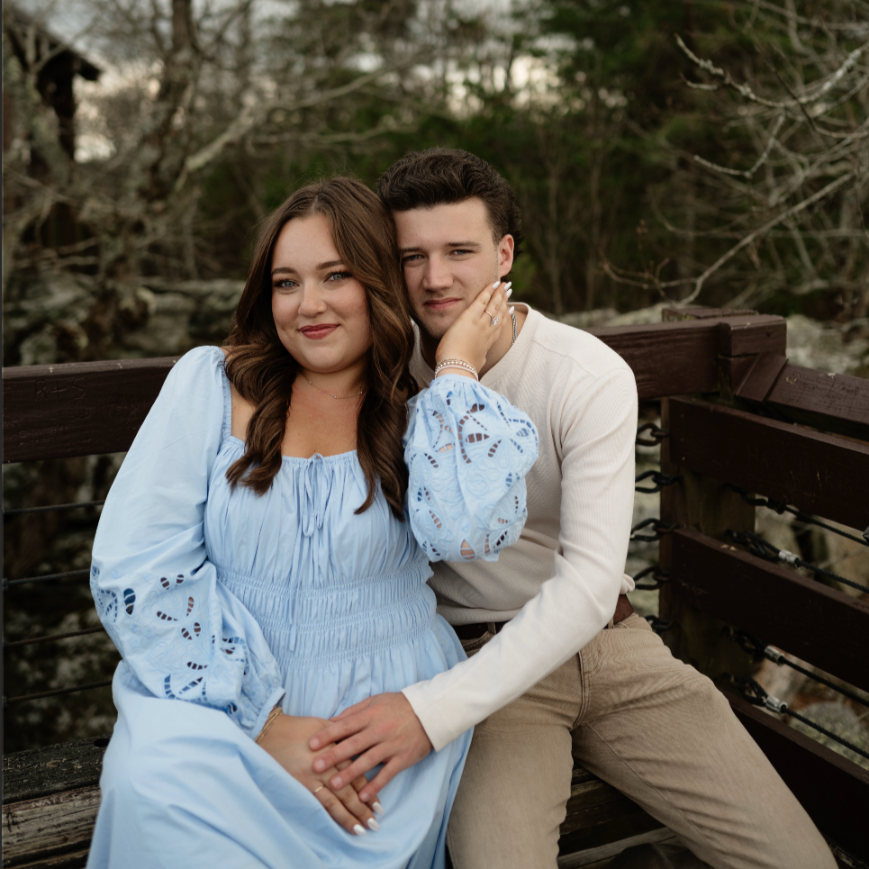 A young couple sitting on a wooden bridge outdoors, with trees and a stream in the background. The woman is wearing a light blue dress, and the man is in a cream sweater and khaki pants. They are close together, with the woman gently touching the man's face and both smiling softly.