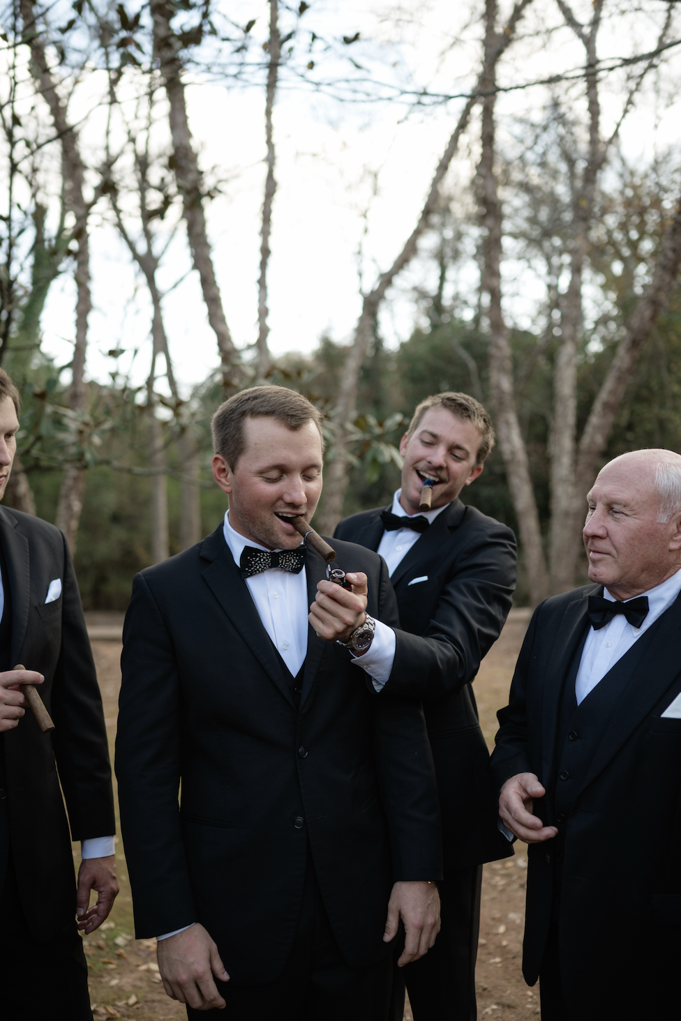 Group of men in tuxedos outside, two smoking cigars and one lighting a cigar.