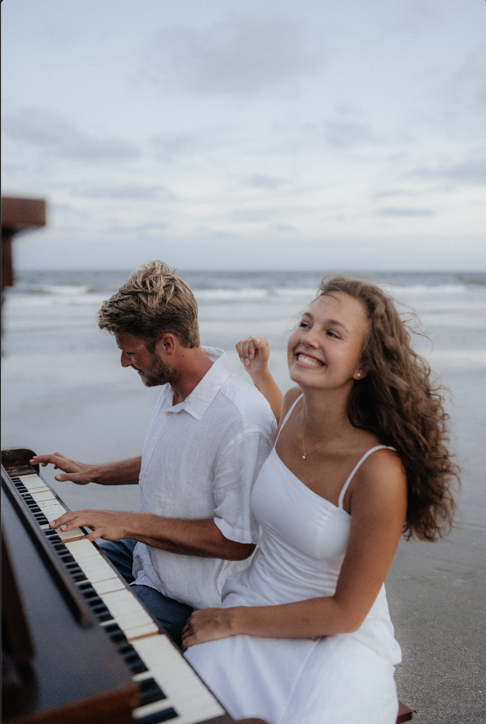 A young woman in a white dress smiling happily while sitting beside a man playing piano on the beach during daytime.