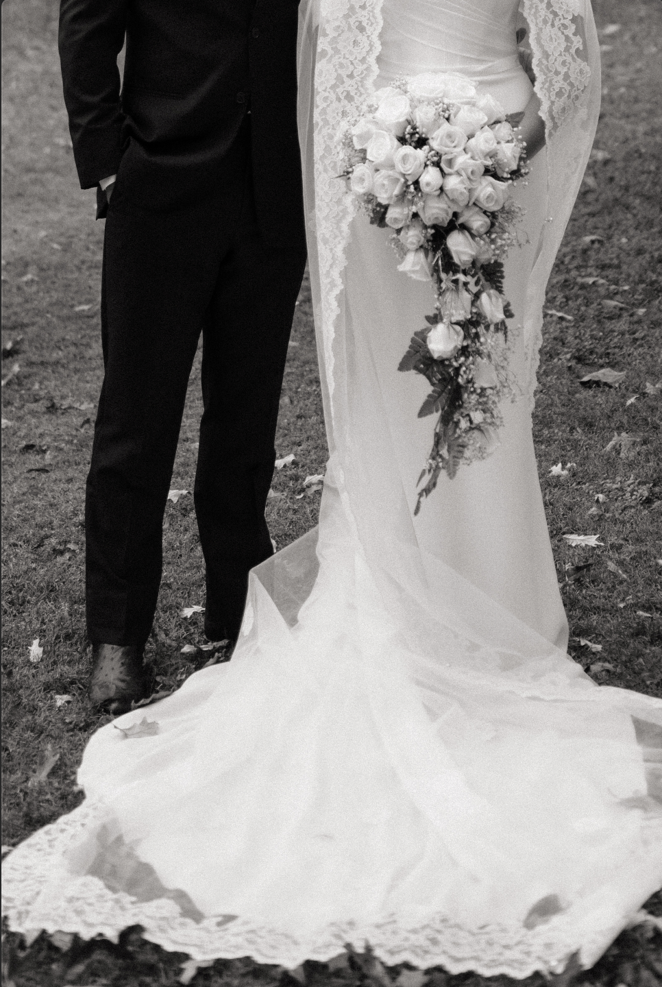 A black and white photo showing a bride and groom standing outdoors on grass. The groom is wearing a dark suit with his hands in his pockets, and the bride is holding a cascading bouquet of roses and greenery, wearing a lace wedding dress with a long