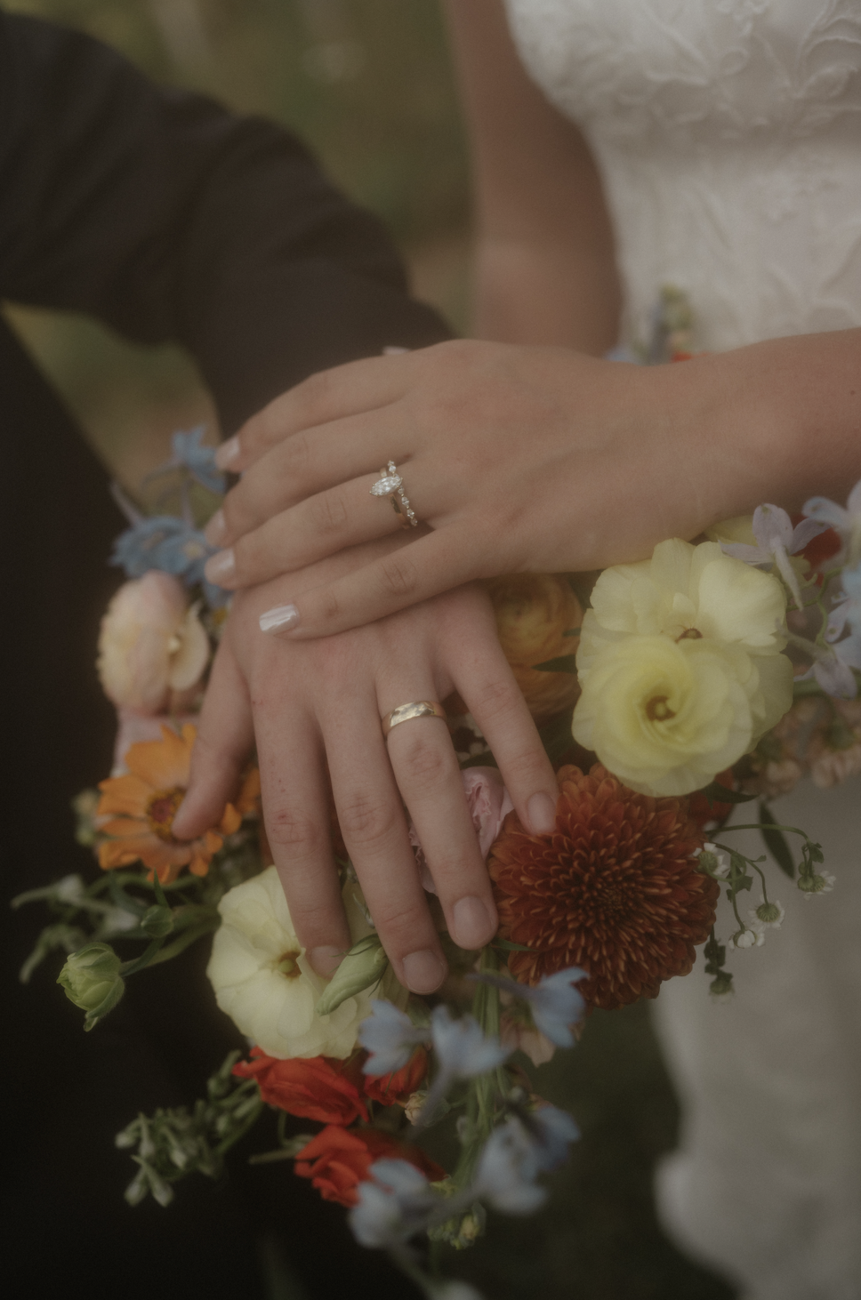 Close-up of a bride and groom's hands showing wedding rings, surrounded by a colorful bouquet of flowers.