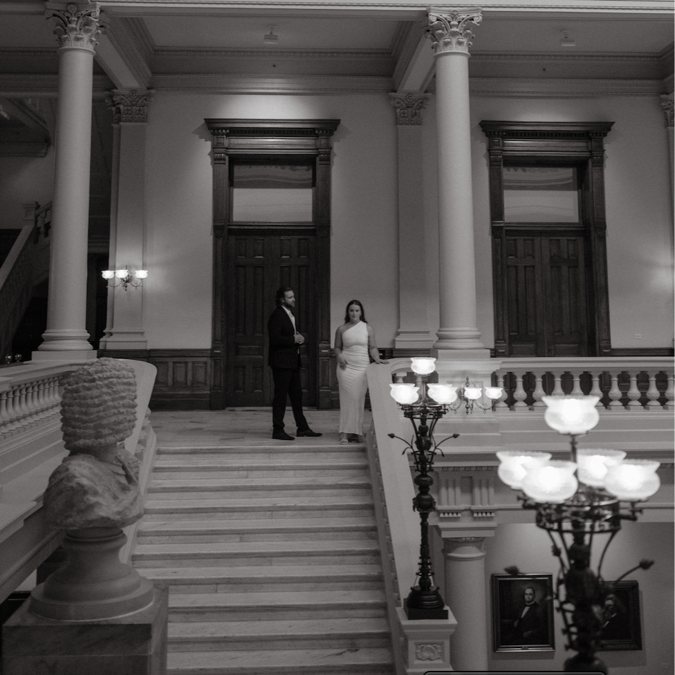A black and white photo of an elegant, historic interior with grand staircase, tall columns, and ornate chandeliers. Two people, a man in a dark suit and a woman in a long gown, stand on the staircase.