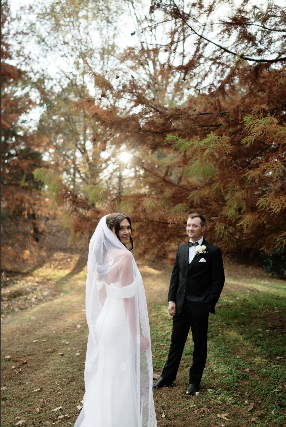 A bride and groom stand outdoors among autumn trees, with the bride wearing a white wedding dress and veil, and the groom in a black tuxedo with a white shirt and bowtie, during a wedding photoshoot.