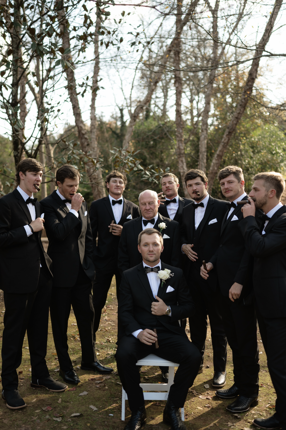 A group of nine men in black tuxedos with bow ties, some holding cigars, gathered outdoors in a wooded area during daylight, with one man seated in front and the others standing behind him.