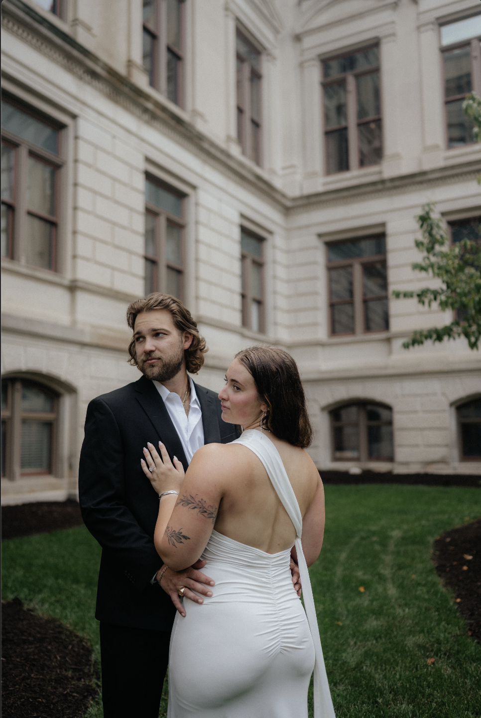 A couple dressed in formal attire stands outdoors in front of a historic building with large windows and cream-colored walls. The woman in a white gown with an open back and a tattoo on her arm leans on the man in a black suit, holding his lapel, as 