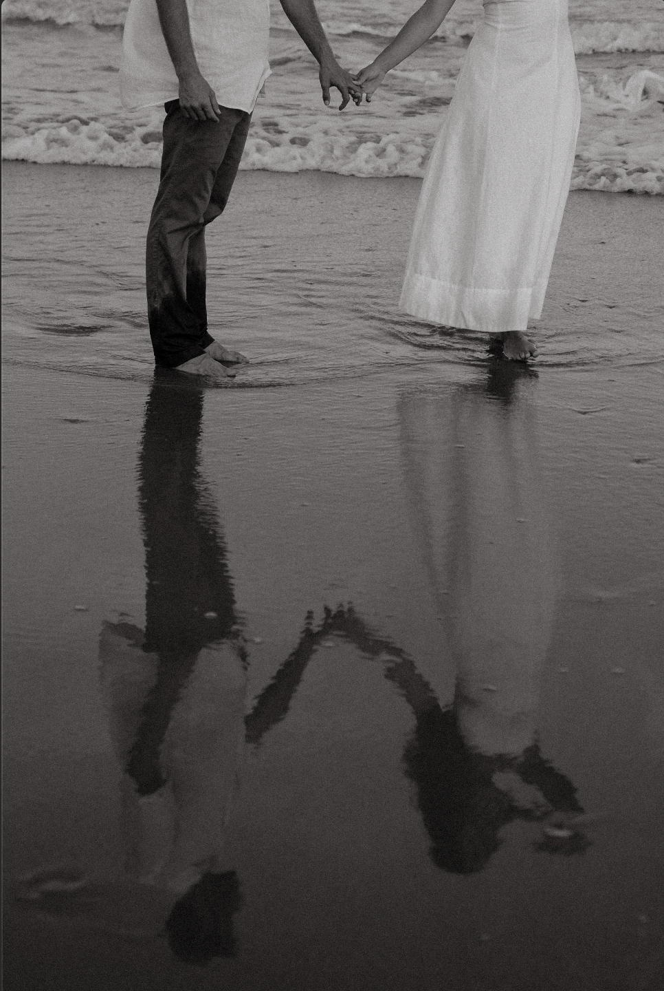 A couple holding hands and walking on the beach, with their reflections visible in the wet sand, in black and white.