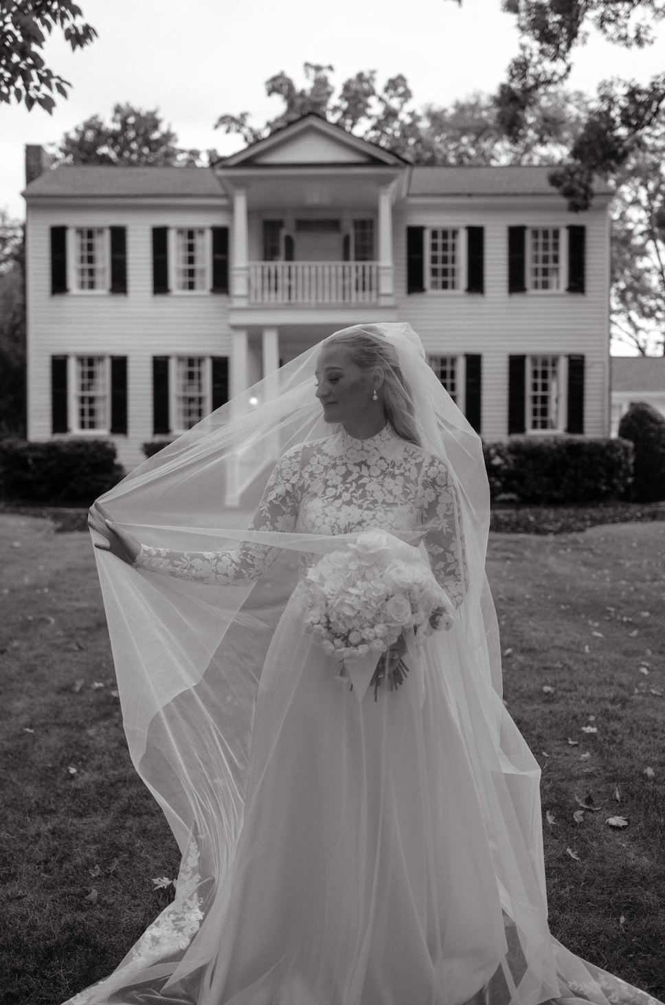 Black and white photo of a bride holding a bouquet, standing outdoors in front of a large house with multiple windows and shutters, her veil flowing over her face and dress.