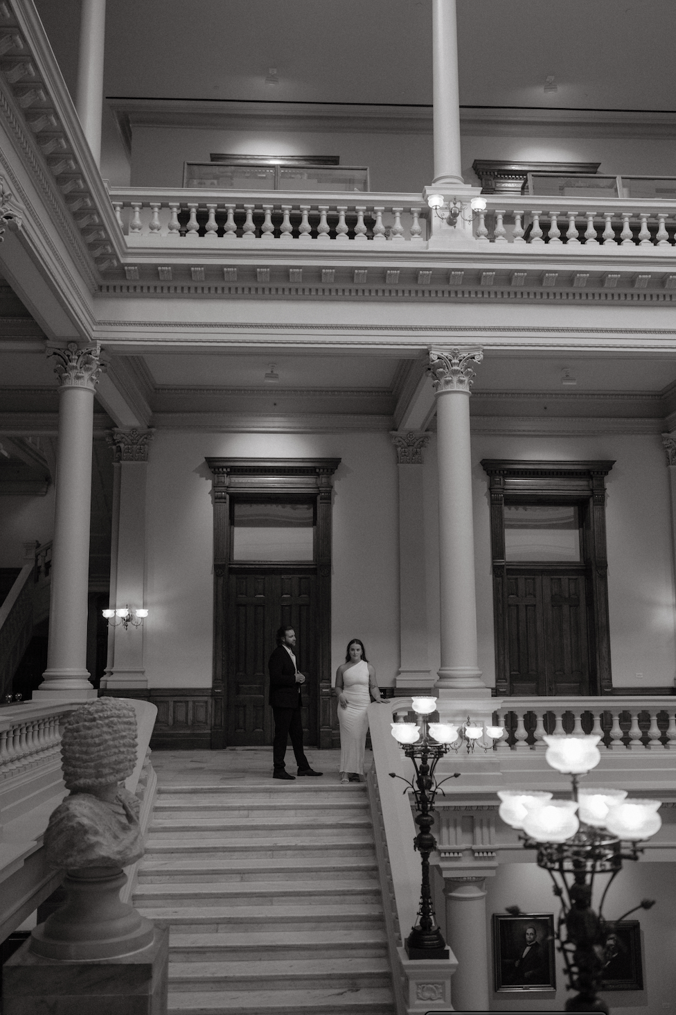A grand, historic interior with marble staircase, high ceilings, ornate columns, and wooden accents; two people in formal attire standing on the stairs, engaging in conversation.