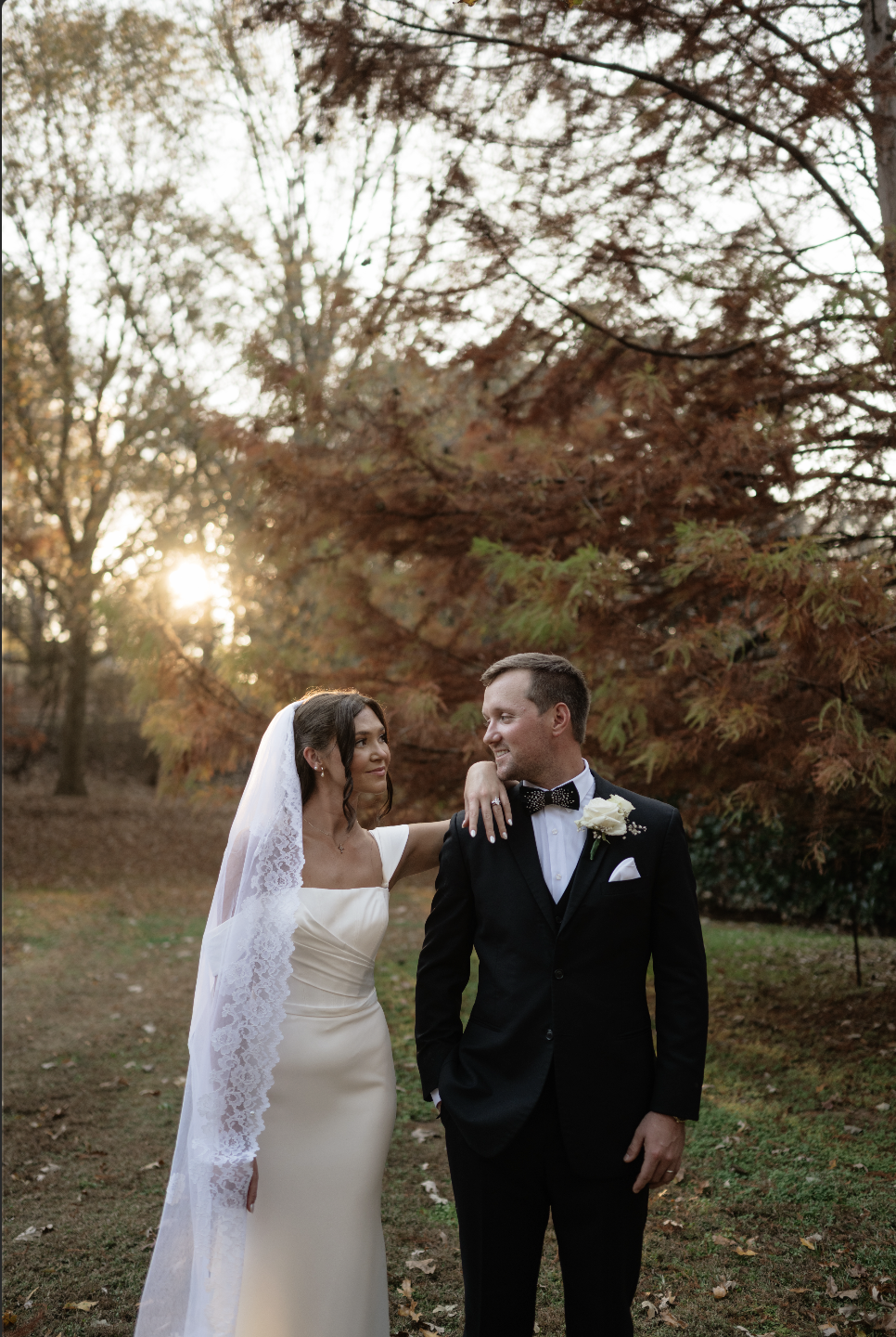 Bride and groom in formal wedding attire standing outdoors among trees with autumn leaves, during sunset.