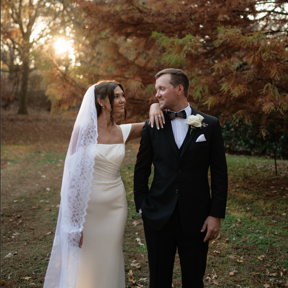 A bride and groom standing outdoors during sunset, smiling at each other with autumn trees in the background.