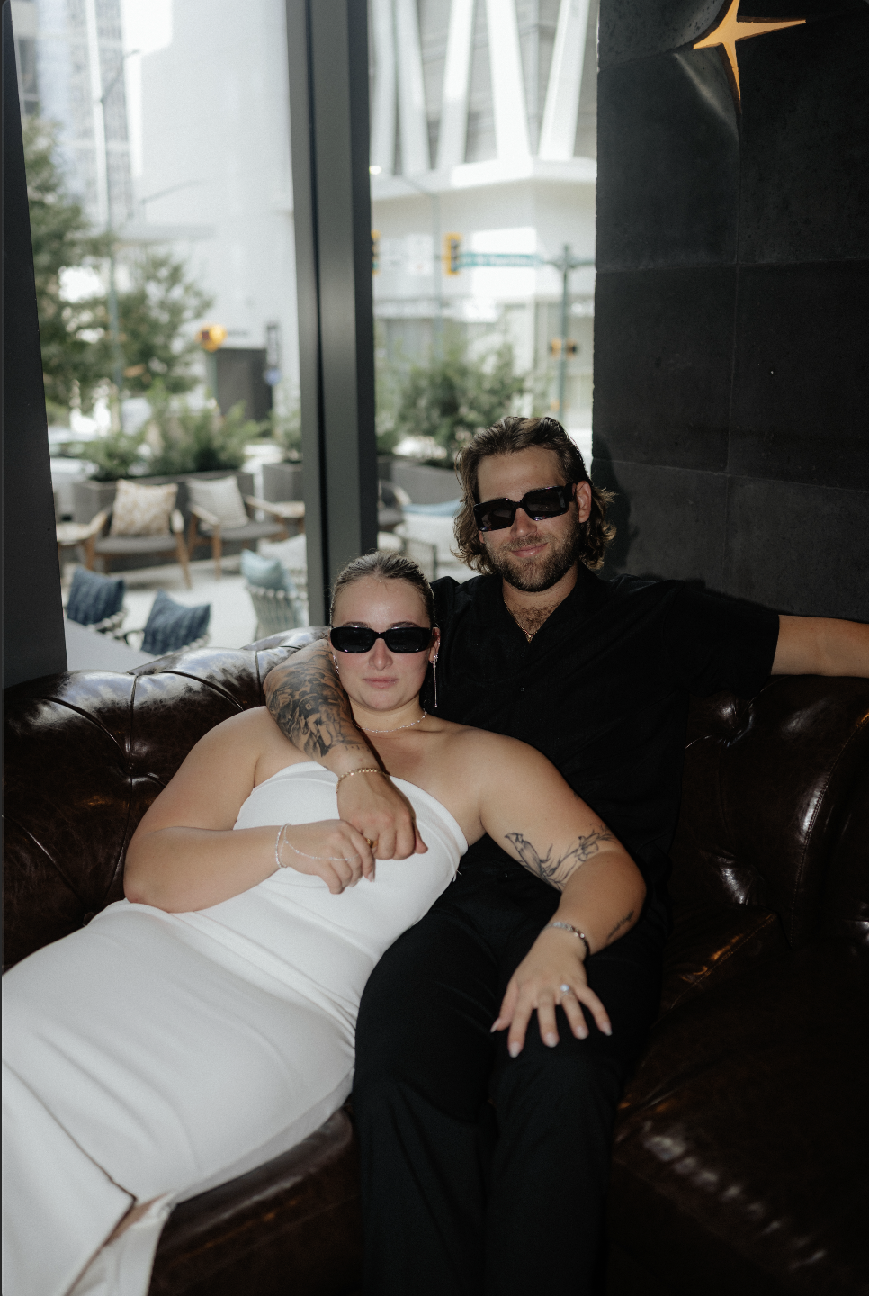 A man and woman sitting on a leather couch, both wearing sunglasses, with the man arm around the woman. The woman is in a white strapless dress, and the man is in a black shirt.