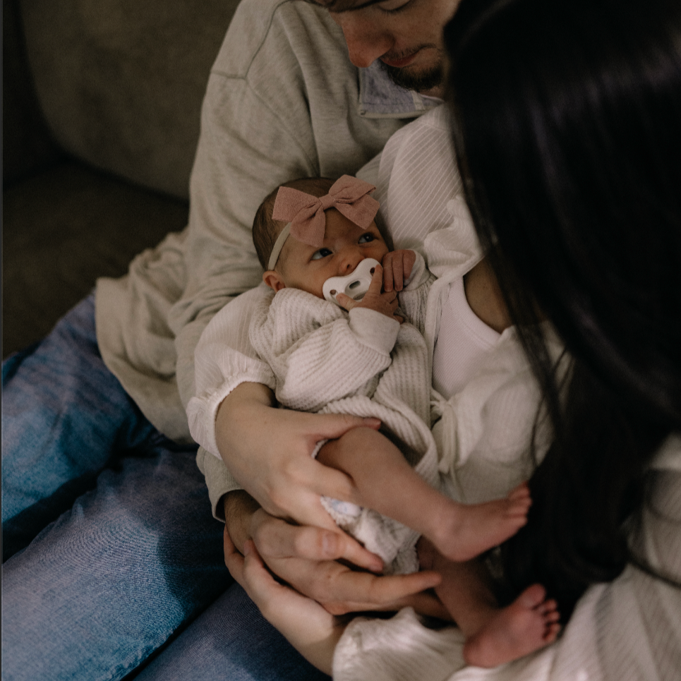 A close-up of a family scene where a baby girl is being cradled by her parents on a couch. The baby is wearing a pink bow headband and a white pacifier, looking curiously at her mother while sucking her finger. The mother, with long dark hair, is holding the baby and looking affectionately at her. The father, with a beard, is leaning in closely from behind, also focused on the baby.