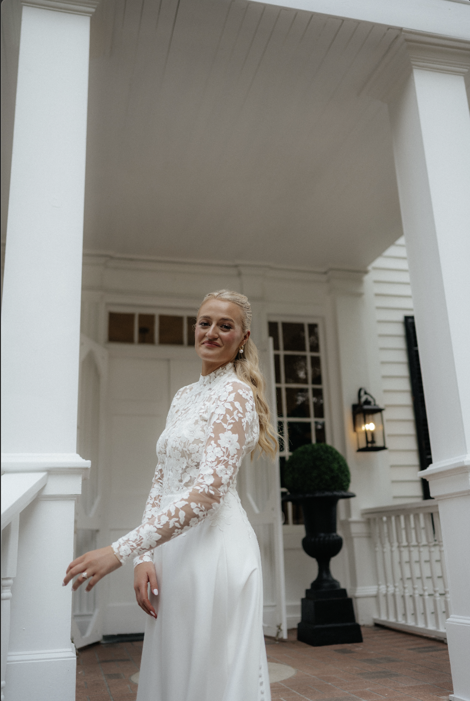 A bride in a white wedding dress with lace sleeves standing on the porch of a white house, smiling at the camera.