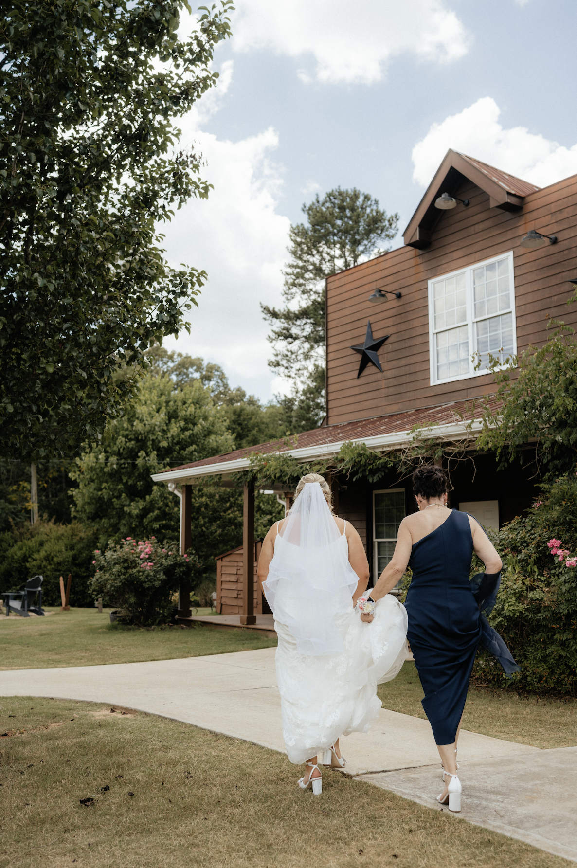 Two women, one in a wedding dress and the other in a blue dress, walk together on a sidewalk in front of a rustic house with a star decoration on the wall, surrounded by trees and flowers.
