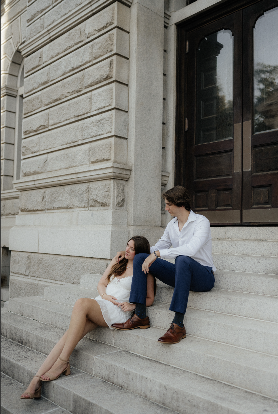 A young man and woman sitting on the steps of a stone building, with a large wooden door behind them. The woman is resting with her eyes closed, wearing a white dress and beige heels. The man is sitting above her, with one leg on the step and the other on the ground, wearing a white shirt, blue pants, and brown shoes, looking down at her.
