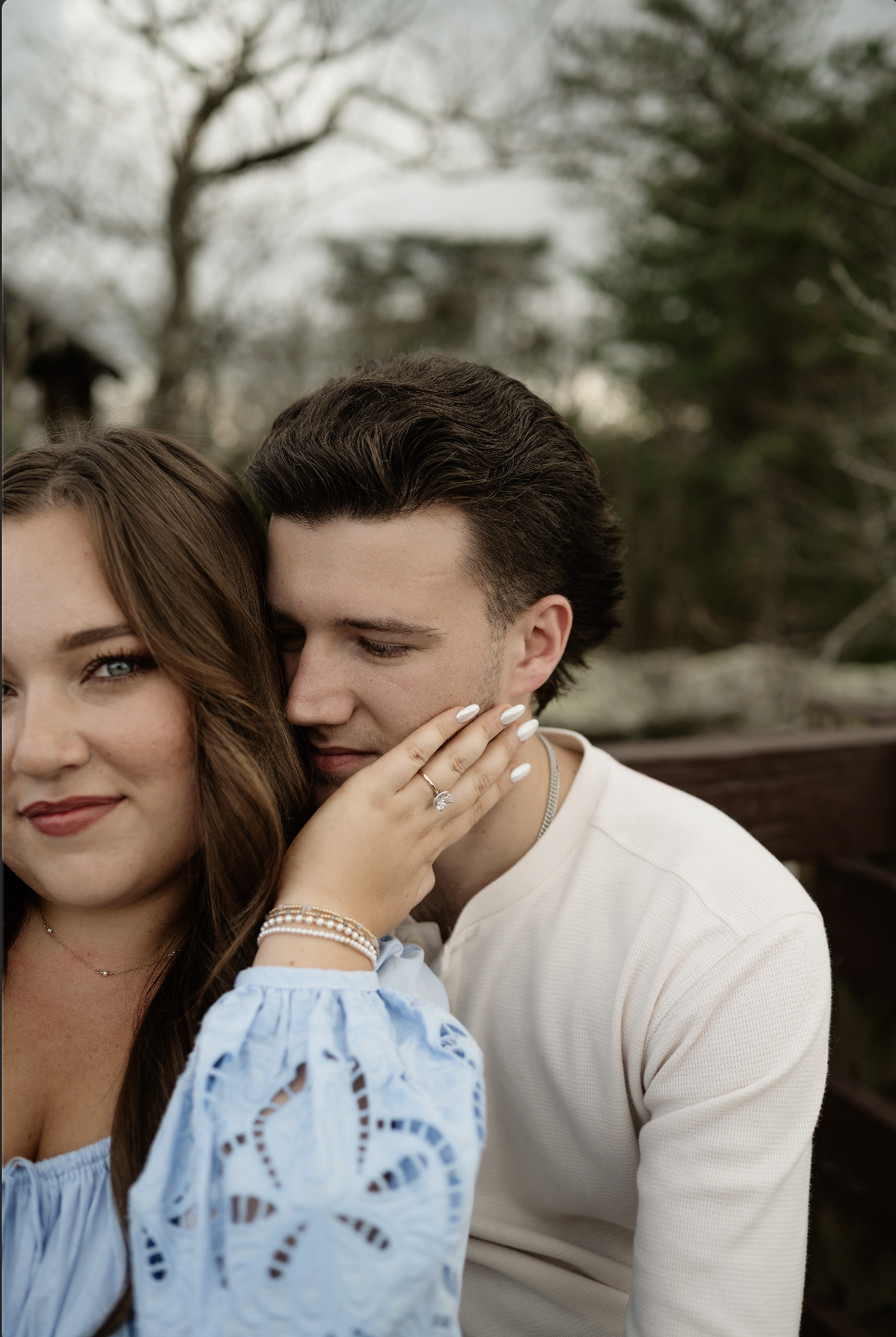 A woman with long, brown hair and blue eyes gently touching a young man's face. The woman is wearing a light blue blouse with lace details, jewelry, and an engagement ring. The young man has short, dark hair and is wearing a light cream-colored shirt